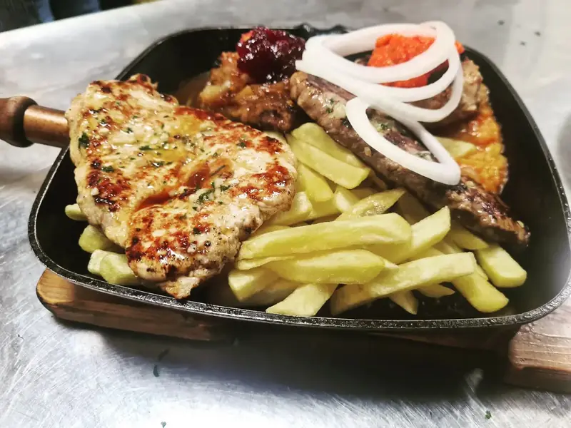 Fleischgericht vom Grill mit Pommes frites, Zwiebelringen, Ajvar und Preiselbeeren in der Pfanne serviert